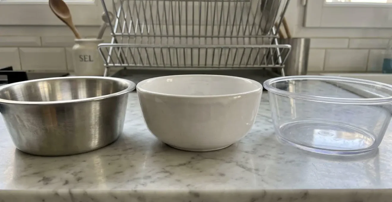 Trois gamelles pour chat alignées sur un plan de travail en marbre blanc moderne : une en acier inoxydable brossé à gauche, une en céramique blanche mate au centre et une en plastique transparent à droite